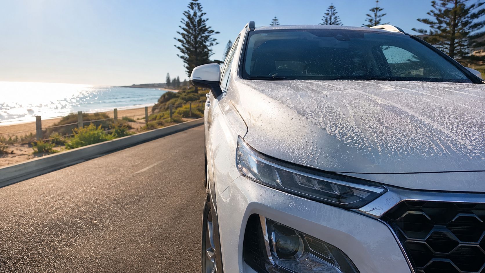 A close-up view of a white car covered in protective film, parked next to a coastal road.