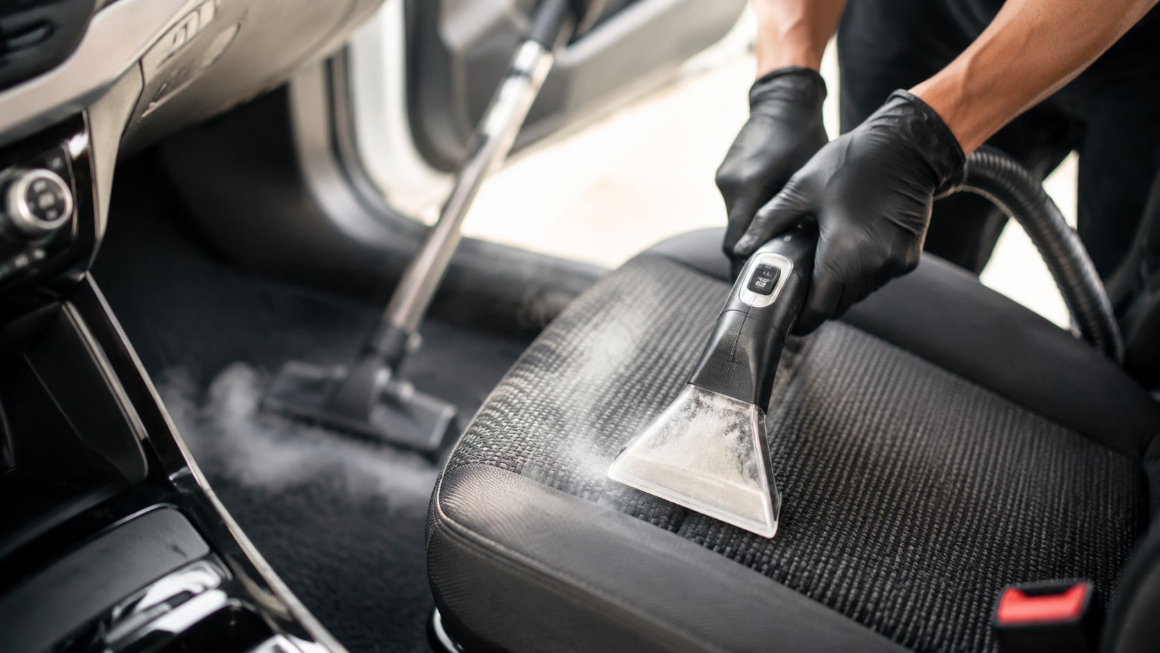 A professional car detailer using a steam cleaner vacuum on the fabric upholstery of a car seat.